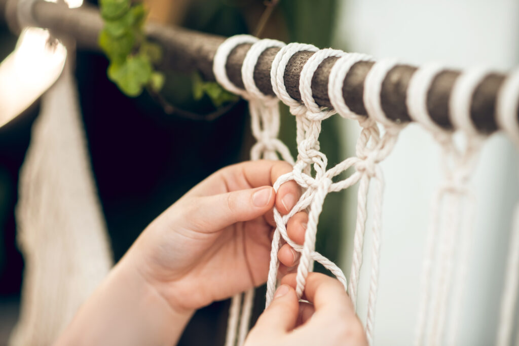 Close-up demonstration of hands tying square knots and spiral knots in white cotton cord for macramé plant hanger construction